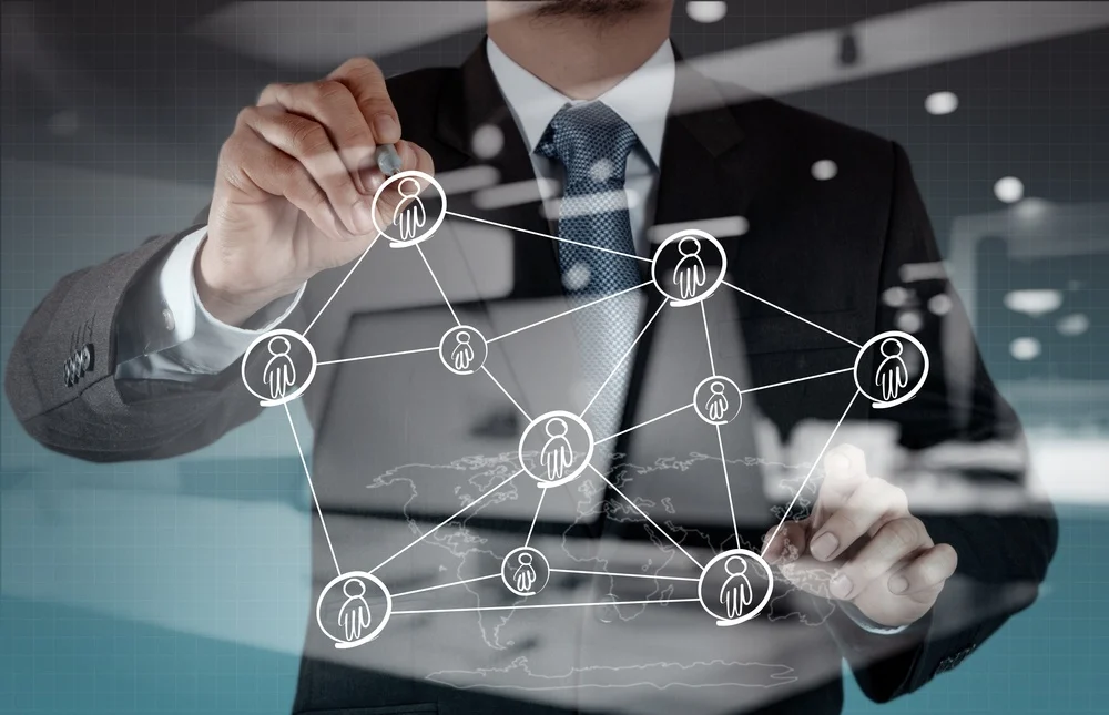 A businessman in a suit draws a virtual network of connected people icons on a transparent screen, symbolizing teamwork, communication, or social networking in a modern office setting.