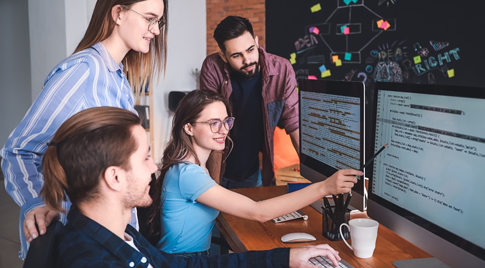 Four people collaborate at a desk with two large monitors displaying code. One person points at the screen while others watch and discuss. Coffee mugs and notebooks are on the desk; a blackboard with sticky notes is in the background.