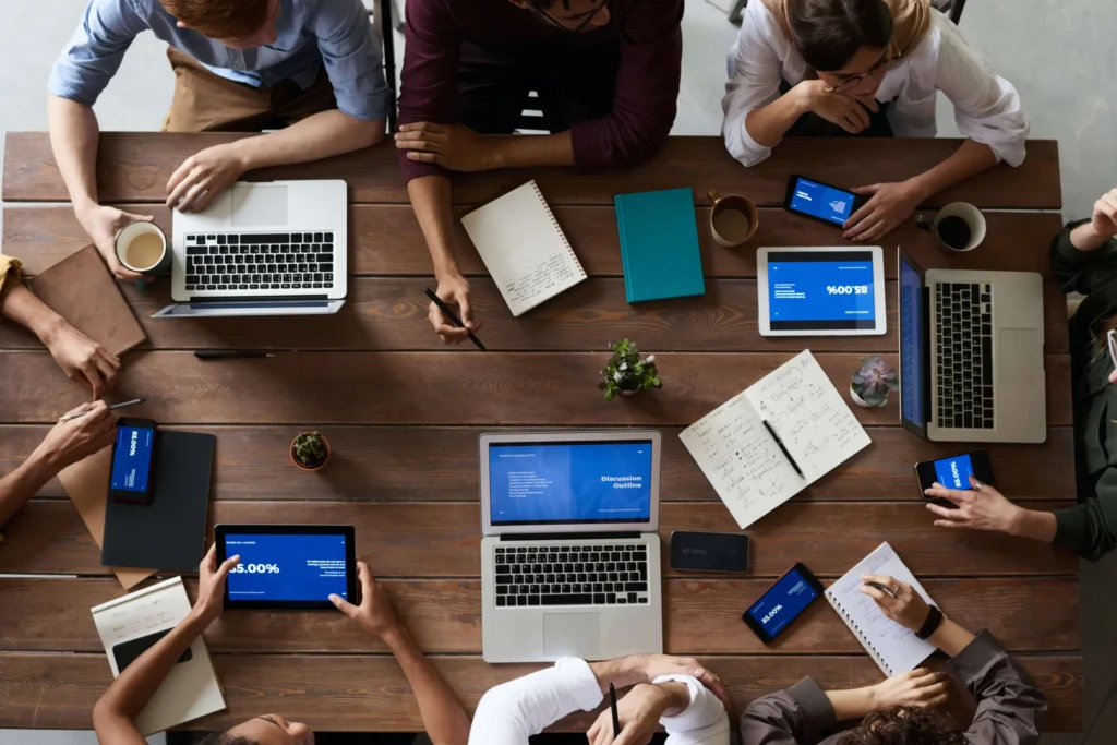 People working together around a wooden table with laptops, tablets, notebooks, and smartphones. Various charts and graphs are visible on the screens, and some people are taking notes. A small plant sits in the center of the table.